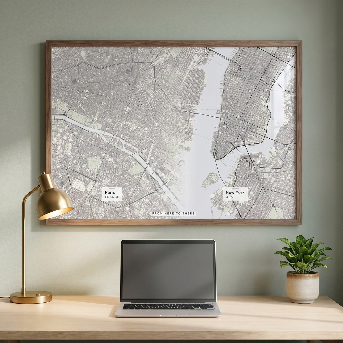 A framed city map wall art print hangs above a light wood desk in a styled room setting, with a gold desk lamp on the left, an open laptop in the center, and a small potted plant on the right.